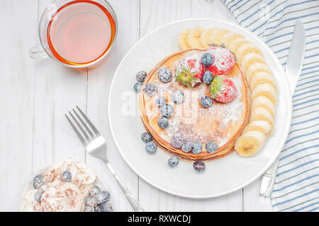 Gesundes Frühstück, schwarzer Tee und hausgemachte Pfannkuchen mit frischen Beeren und Bananen auf Holz Tisch. Stockfoto