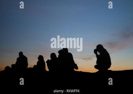 Silhouetten der sitzenden Menschen am Boden in der Dämmerung, von hinten während der schönen Sonnenuntergang fotografiert. Stockfoto