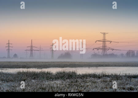 Hohe Spannung elektrische Pole. Elektrische powerline in der Natur. Morgen Nebel unter den Sonnenaufgang Himmel. Stockfoto