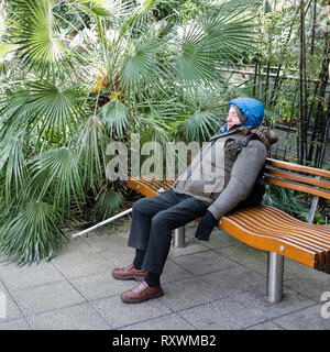 Ein älterer Mann mit einer Gehhilfe schlafend auf einer Bank in einer tropischen Umgebung. Stockfoto