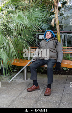 Ein älterer Mann mit einer Gehhilfe schlafend auf einer Bank in einer tropischen Umgebung. Stockfoto