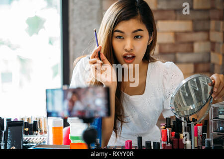 Hübsche asiatische Frau Aufnahme Makeup Tutorial Video über Kosmetika mit Handy auf Stativ Stockfoto