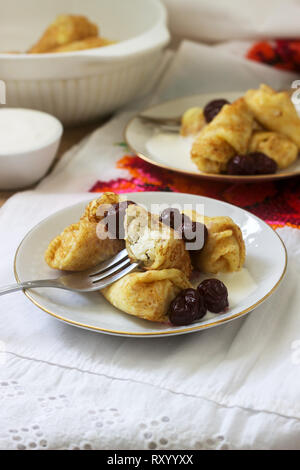 Hausgemachte Pfannkuchen gefüllt mit Hüttenkäse mit Rosinen, serviert mit saurer Sahne und Kirschen. Traditionelle moldawische oder rumänischen Dessert, selektive fo Stockfoto