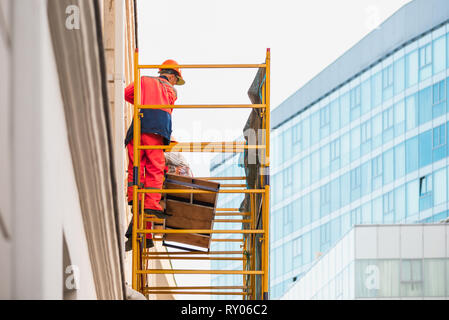 Builder in orange Robe und Schutzhelm steht auf gelb Gerüst lehnte sich gegen das Gebäude vor dem Hintergrund eines modernen Blau Stockfoto