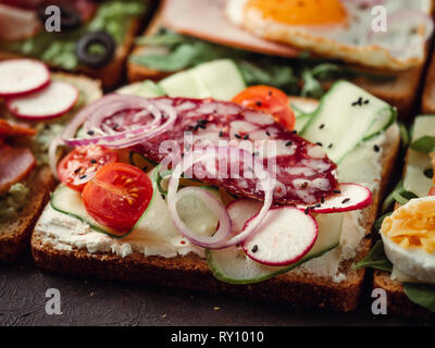 Nahaufnahme der verschiedenen Sandwiches mit Salami, Gemüse und schwarzem Sesam. Kopieren Sie Platz für Text. Sortiment Fleisch Toast auf schwarzem Hintergrund. Idee, kreatives Konzept für Wurst. Stockfoto