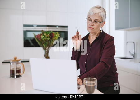 Porträt eines reifen älteren Frau Brille arbeitet sie an ihrem Laptop in der Küche, Denken und auf den Bildschirm mit dem Stift in Ihrer Hand Stockfoto