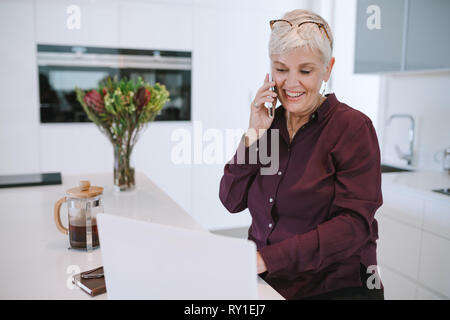 Lächelnd casual ältere Frau mit Laptop während des Sprechens auf dem Smartphone. Gerne reife Frau mit Handy und Laptop zu Hause arbeiten. Business woman u Stockfoto