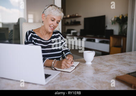 Reife Frau, schön Notizen zu machen, während auf Ihrem Laptop zu Hause arbeiten. Ältere Frau schreiben in Ihrem Notebook draußen auf dem Balkon Stockfoto
