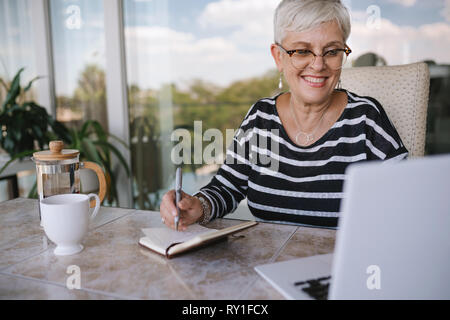 Schöne ältere Frau mit Laptop zu Hause. Reifen lächelnde Frau auf der Suche nach Bildschirm, während das Schreiben im Notebook. Portrait von glücklich Moderne pensionierten Dame w Stockfoto