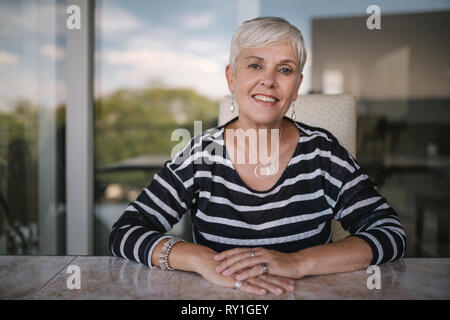 Schöne ältere Frau lächelnd in die Kamera. Porträt einer reifen Frau mit Händen auf dem Tisch gekreuzt, draußen auf dem Balkon. Schöne Stockfoto