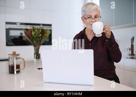 Schöne ältere Frau mit Laptop Computer zu Hause, im Internet surfen. Ältere Dame, indem Sie auf Computer und genießen eine Tasse Kaffee Stockfoto