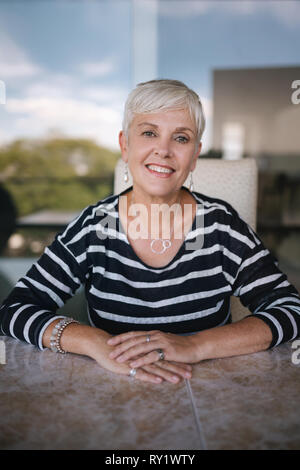 Schöne ältere Frau lächelnd in die Kamera. Porträt einer reifen Frau mit Händen auf dem Tisch gekreuzt, draußen auf dem Balkon. Schöne Stockfoto