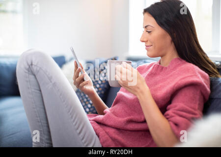 Junge Frau auf Sofa mit heißen Getränken Kontrolle Mobiltelefon Stockfoto