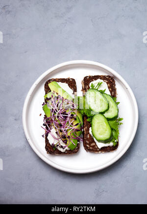 Roggen Brot mit Avocado, Sahne, Käse, Gurken, Sprossen in eine weiße Platte auf grauem Hintergrund Stockfoto