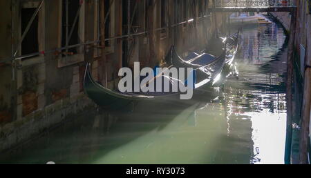 Sonnenstrahlen, die durch die kleine Lücke der Gebäude auf dem Wasser Kanal in Venedig mit den drei Gondeln Docking an der Seite in Venedig Es widerspiegelt Stockfoto