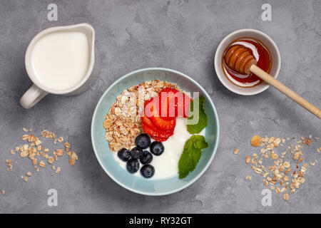 Gesundes Frühstück Müsli mit Joghurt, Erdbeeren und Blaubeeren in einem blauen Schale auf einem grauen Hintergrund. Ansicht von oben Stockfoto