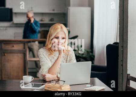 Selektiver Fokus der nachdenkliche Frau, die in der Nähe von Laptop mit Ehemann auf Hintergrund Stockfoto