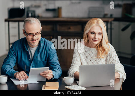 Schöner Mann in Gläsern mit digitalen Tablet in der Nähe der attraktiven Blondine Frau an Laptop suchen Stockfoto