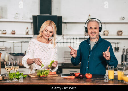 Fröhliche Frau die Zubereitung von Speisen in der Nähe von Happy Mann Musik hören über Kopfhörer in der Küche Stockfoto