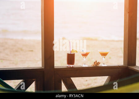 Frische exotische Cocktails auf hölzernen Rand. Shell liegt zwischen Brille. Cola mit Strohhalm und Zitrone. Flanke von Haus. Blick auf das Meer. Stockfoto