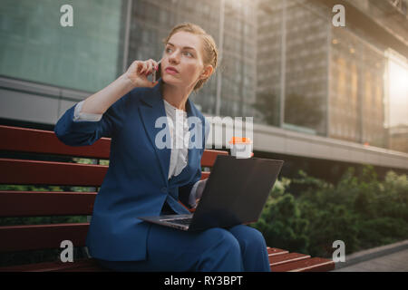 Junge Frau sitzt im Freien und mit Laptop. Geschäftsfrau. Die Tasse Kaffee. Und sprechen gleichzeitig am Telefon. Stockfoto