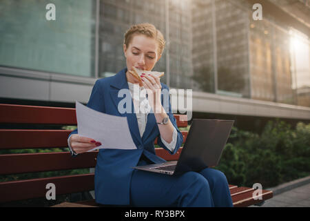 Beschäftigte Frau ist in Eile, sie hat keine Zeit, sie wird Imbiss im Freien zu essen. Arbeitnehmer essen und Arbeiten mit Dokumenten auf dem Laptop an der Stockfoto