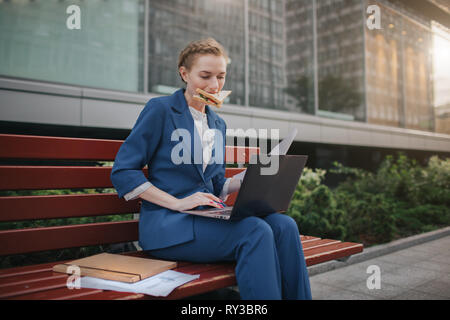 Beschäftigte Frau ist in Eile, sie hat keine Zeit, sie wird Imbiss im Freien zu essen. Arbeitnehmer essen und Arbeiten mit Dokumenten auf dem Laptop an der Stockfoto