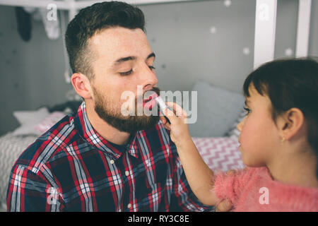 Süße kleine Tochter und ihrem stattlichen jungen Eltern sind zusammen spielen im Kinderzimmer. Mädchen tut ihr Vati ein Make-up. Stockfoto