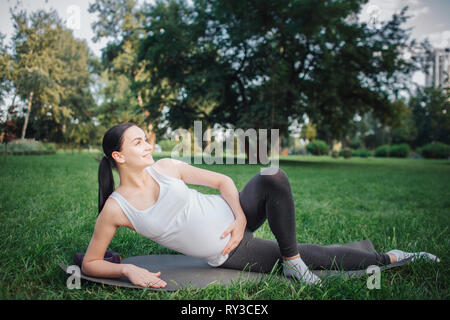 Freundliche nette junge schwangere Frau liegen auf der einen Seite und außerhalb lächelnd in Park. Sie ein anderes halten sie auf dem Bauch. Das linke Bein vor der Rechten. Stockfoto