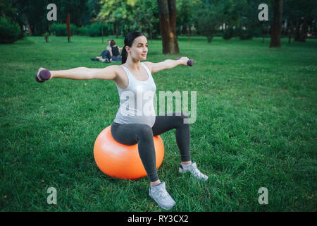 Gut gebauten jungen schwangeren Frau auf orange Fitness Ball sitzen und das Trainieren im Park. Sie halten Hanteln. Stockfoto