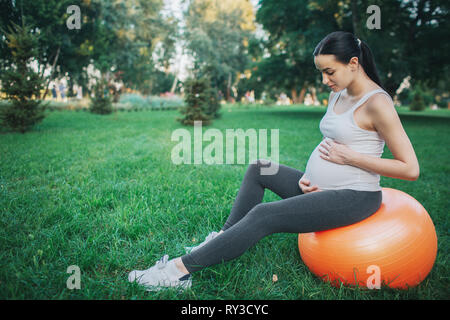 Hübsche junge schwangere Frau sitzen auf orange Fitness Ball im Park. Sie halten Hände um Bauch und schauen Sie es. Stockfoto