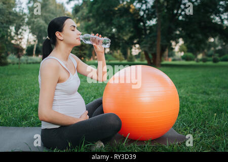 Nette junge oregnant Frau trinkt Wasser im Park. Sie sitzen auf Yoga mate und eine Hand auf dem Bauch halten. Orange Fitness Ball neben liegen. Stockfoto