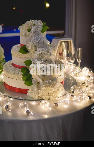 Ein 3-tiers weisse Hochzeitstorte mit rose Dekore auf eine Tabelle mit weissen Kerzen und Lampen eingerichtet. Hochformat. Stockfoto
