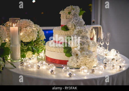 Ein 3-tiers weisse Hochzeitstorte mit rose Dekore auf eine Tabelle mit weissen Kerzen und Lampen eingerichtet. Querformat. Stockfoto