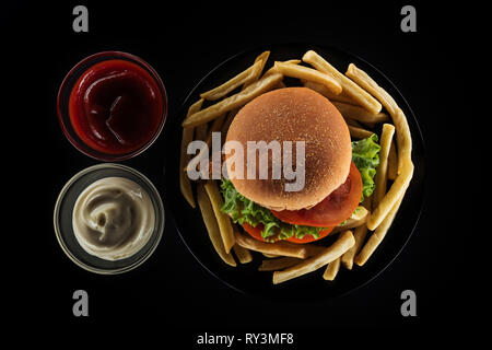 Blick von oben auf die leckere Pommes frites und Chicken Burger in der Nähe von Mayonnaise und Ketchup isoliert auf Schwarz Stockfoto