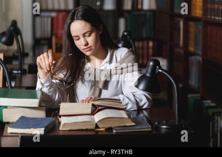 Frustriert, Studentin am Schreibtisch sitzen und mit einem riesigen Haufen von Studie Bücher in der Universitätsbibliothek Stockfoto