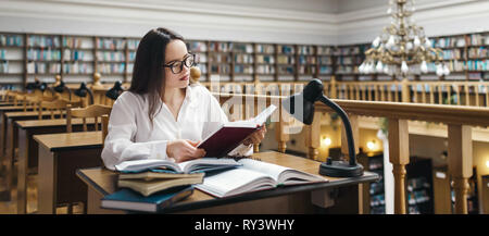 Frustriert, Studentin am Schreibtisch sitzen und mit einem riesigen Haufen von Studie Bücher in der Universitätsbibliothek und Studieren Stockfoto