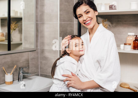 Tochter und Mutter in weißen Bademänteln umarmen im Badezimmer Stockfoto
