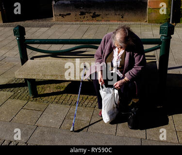 Eine ältere Frau sitzt auf der öffentlichen Bank suchen in Shopping Bag genießen Sie sonnige Wetter in Fort William High Street lochaber Stockfoto