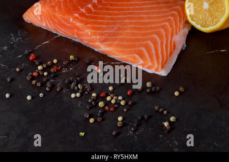 Frischer Lachs in der Platte mit Zitronenscheiben ang Herden auf dunklem Hintergrund. Organische Zutaten zum Kochen für Meeresfrüchte Stockfoto