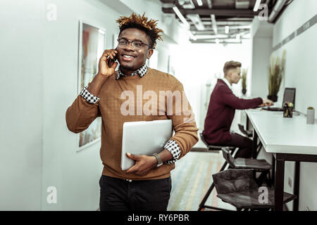 Strahlende Kerl in klare Gläser mit Dreadlocks in Konversation auf dem Smartphone Stockfoto