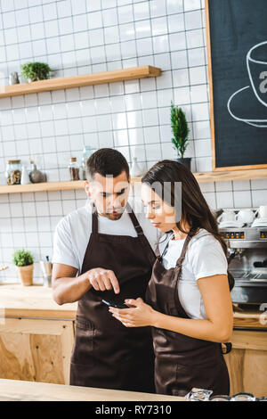 Kassierer in Braun Schürzen stehen hinter Theke und Suchen in Coffee House auf Smartphone Stockfoto