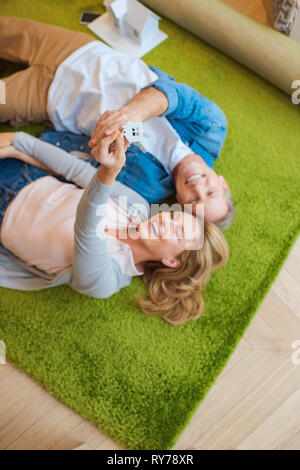 Selektiver Fokus der Frau mit Tasten mit Haus Modell Schmuckstück beim Lügen auf grünen Teppich mit lächelnden Mann Stockfoto