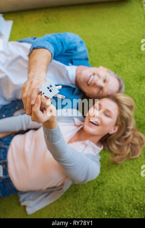 Selektiver Fokus der glücklichen Paar hält Tasten mit Haus Modell Schmuckstück beim Lügen auf grünen Teppich Stockfoto