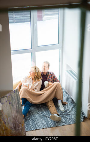 Glückliches Paar sitzen auf dem Boden durch die großen Fenster und halten Kaffeetassen Stockfoto