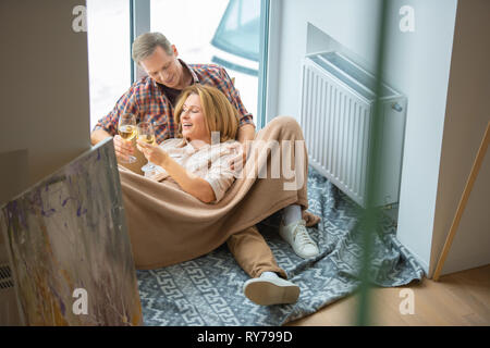 Glückliches Paar anstoßen, während auf dem Boden durch die großen Fenster im neuen Zuhause ruhen Stockfoto