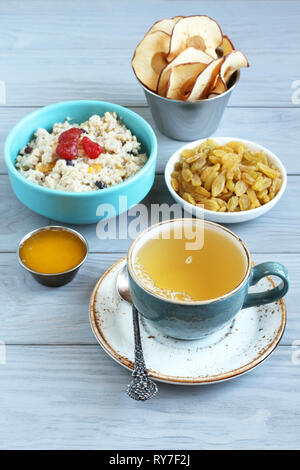 Frühstück: Haferflocken mit Obst Stücke und Kandierte Erdbeeren, weiß Rosinen, Apfel Chips, Honig in kleinen Schüsseln und eine Tasse grünen Tee auf einem grauen Woode Stockfoto