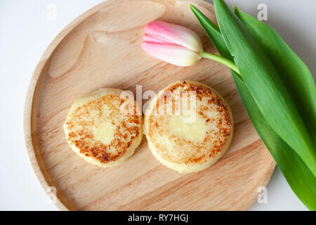 Nahaufnahme von frischen rosa Tulpe auf Holz- runde Platte, zwei Goldene gebissen fried Käsekuchen in der Form der Abbildung 8, Dessert von Hüttenkäse. Con Stockfoto