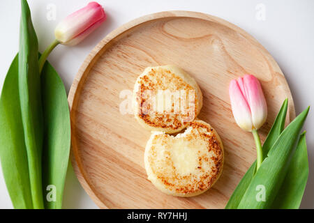 Nahaufnahme von frischen rosa Tulpe auf Holz- runde Platte, zwei Goldene gebissen fried Käsekuchen in der Form der Abbildung 8, Dessert von Hüttenkäse. Con Stockfoto