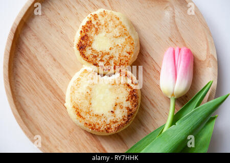 Nahaufnahme von frischen rosa Tulpe auf Holz- runde Platte, zwei Goldene gebissen fried Käsekuchen in der Form der Abbildung 8, Dessert von Hüttenkäse. Con Stockfoto